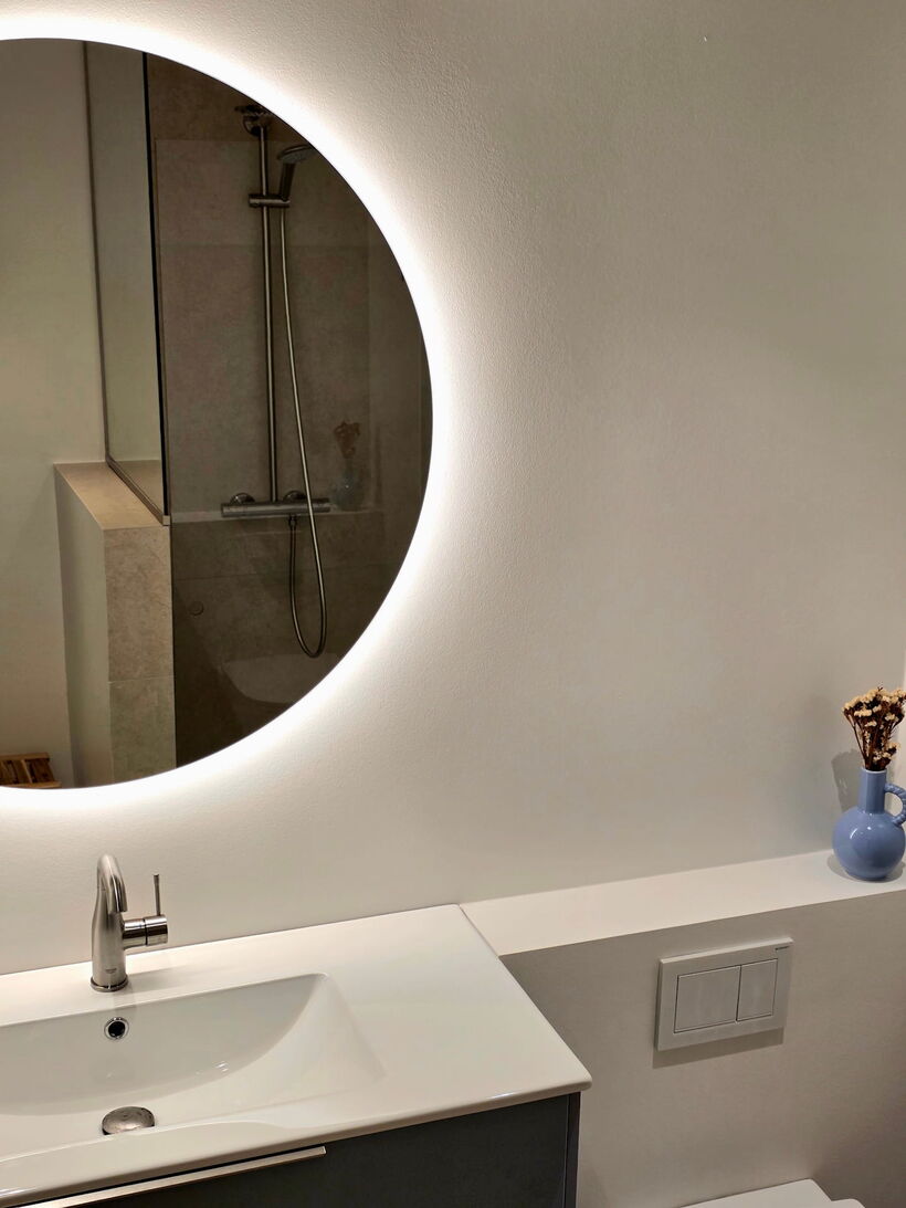 Renoveret badeværelse i Charlottenlund med rundt baglyst spejl, porcelænsvask og walk-in bruser i naturstensfliser, udført af Fokus Bad